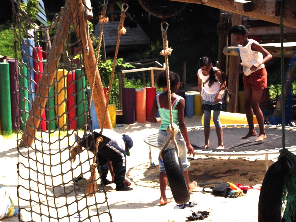 A photo of four children playing on a sanded area on an adventure playground. Multicoloured poles protrude from the ground, as a fence, in the background. A tire swing and cargo netting hang in the foreground. Two children and stood on the sand watching two other children jump on a black trampoline. 