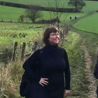 A square photo of Sarah Wilson. She appears to be on a dirt track in open countryside, with green fields, hedgerows, and walkers in the background. Sarah is smiling with her hand on her right hip. She has short dark hair, glasses, and dressed all in black.