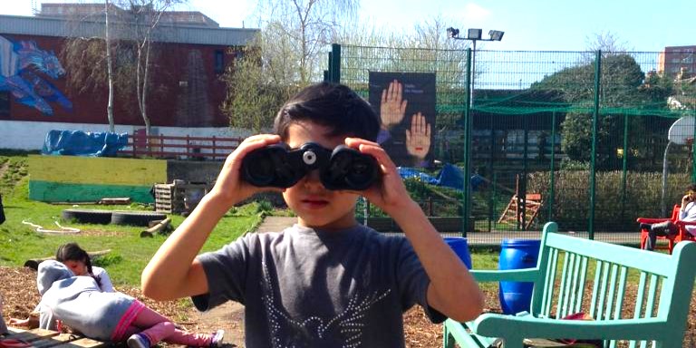 A young boy holds a pair of binoculars up to his eyes and stares at the camera. Two young girls are sat on the floor, behind him and to the left, playing with something unseen on the ground between them. 