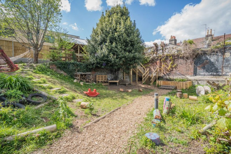 A large tree is in the background with a blue sky with white clouds above. A grassed bank and glimpses of a rope hammock are on the left. On the right a wooden staircase wind up and around the back of the tree to what looks like a covered area with a corrugated roof. 