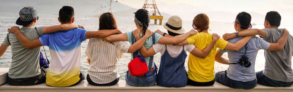 A line of young people are sat with their backs to the camera with their arms around each others shoulders.