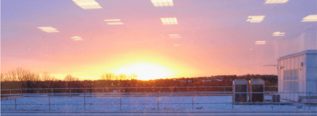 Sunrise view from a building, featuring a vibrant orange and pink sky with silhouettes of trees and a railing in the foreground.